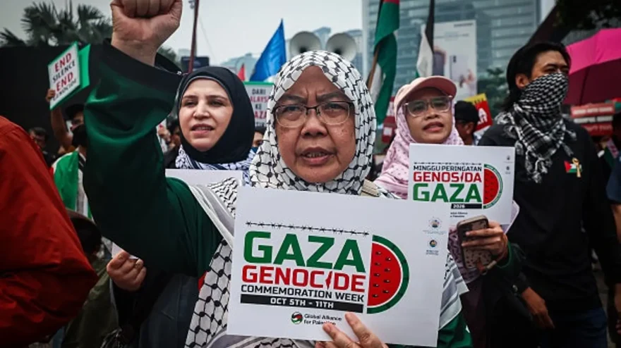 A woman covers her hairs with a Palestinian keffiyeh scarf and raises her fist while holding a placard reading "Gaza Genocide" during an anti-Israel rally Oct. 9, 2026, in Kuala Lumpur, Malaysia. The sign is part of a series of downloadable pre-made graphics created by organizers of the global protest. (Annice Lyn/Getty Images)